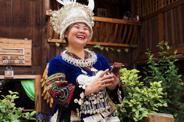 China, Guizhou, laughing Miao woman wearing traditional dress and headdress holding cell phone - obrazy, fototapety, plakaty