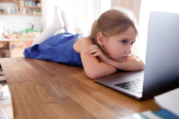 Portrait of sad little girl lying on kitchen table at home looking at laptop