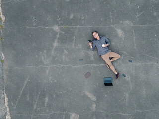 Young businessman with coffee to go lying on tarmac using mini tablet, quadcopter view