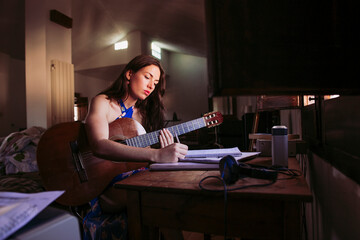Young woman writing in book while practicing guitar at home - obrazy, fototapety, plakaty