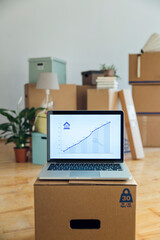 Rising line graph on laptop screen in front of cardboard boxes in an empty room in a new home