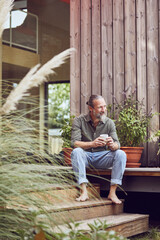 Thoughtful mature man holding coffee cup while sitting outside tiny house