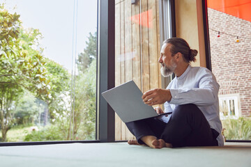 Bearded mature man with laptop looking through window while sitting in tiny house