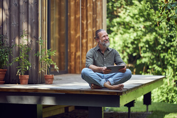 Bearded mature man holding digital tablet contemplating while sitting outside tiny house