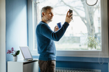 Mature man with coffee mug standing in living room taking selfie with smartphone