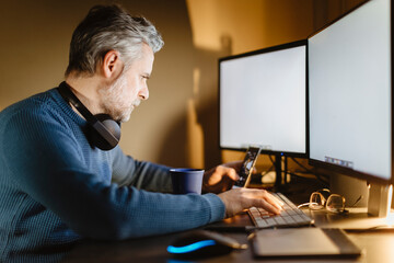 Mature man sitting at desk at home working with smartphone and computer