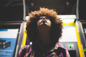 Young woman with afro hairdo at ticket machine at night looking up