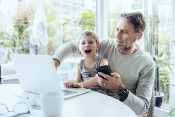 Little boy sitting on lap of father, traying to work from home