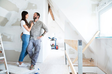 Smiling couple standing in attic to be renovated - obrazy, fototapety, plakaty