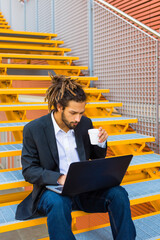 Young businessman with dreadlocks sitting on stairs using laptop