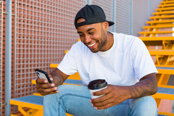 Laughing young man with coffee to go sitting on stairs looking at smartphone