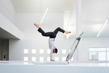 Businessman doing a handstand at laptop in office - obrazy, fototapety, plakaty