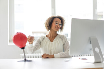 Portrait of young freelancer sitting at desk in office boxing punching ball while looking at...