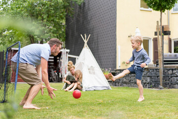 Man and son playing soccer while woman sitting with daughter in tent at back yard during weekend