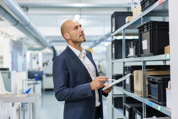 Mature male professional with digital tablet looking at containers on rack in industry