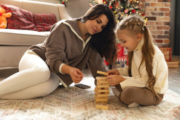 Happy mom and cute little girl are sitting on floor near Xmas tree playing lotto board game...