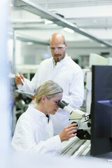 Female scientist doing research while looking into microscope by male colleague in laboratory