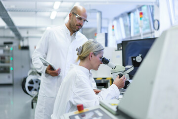 Male technician standing by female scientist looking into microscope while doing research in...