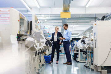 Male technician holding digital tablet while discussing with businesswoman standing by machinery at...