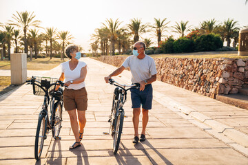 Senior man and woman with face mask talking while walking with cycle at park