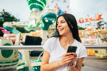 Cheerful young woman holding smart phone looking away at amusement park 