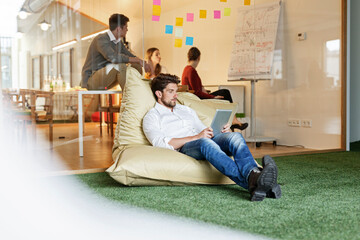 Man in office using tablet in bean bag with meeting in background