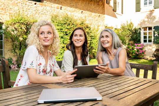 Three women of different age sitting at garden table using tablet