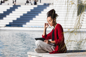 Young woman sitting cross-legged using digital tablet while at edge of promenade over river on...
