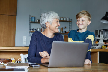 Smiling grandson standing next to grandmother working on laptop at home