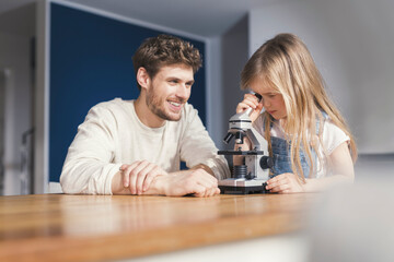 Father watching daughter use a microscope, smiling proudly