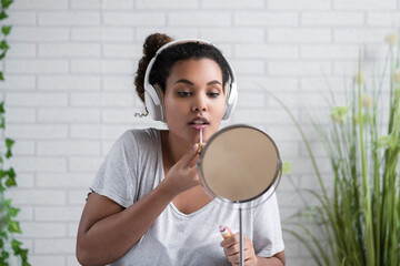 Young woman listening music through headphones applying lip gloss at home