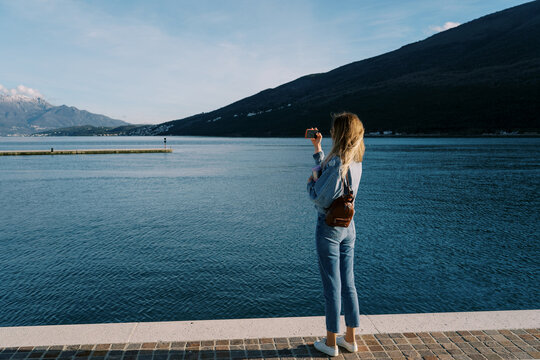 Woman takes pictures of the sea and mountains with a smartphone while standing on the pier. Back view