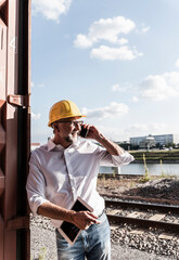 Businessman at cargo harbour, wearing safety helmet, using smartphone and digital tablet