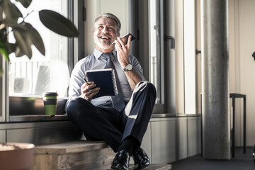 Happy mature businessman with notebook sitting at the window using cell phone