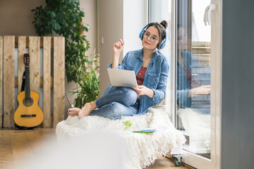 Woman with headphones and laptop sitting at the window at home