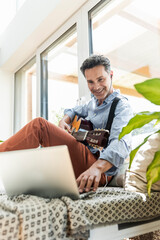 Mature man practising on the guitar, using laptop and earphones