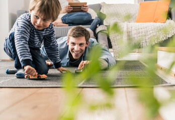 Father and son playing with toy cars, lying on floor - obrazy, fototapety, plakaty
