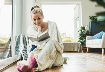 Woman wrapped in a blanket sitting at the window at home reading a book