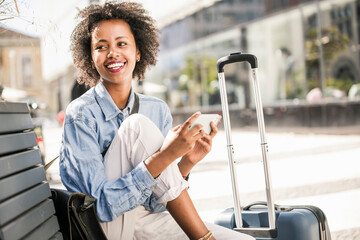Happy young woman sitting on a bench with cell phone and suitcase