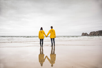 Young woman wearing yellow rain jackets and standing at the beach, Bretagne, France - obrazy, fototapety, plakaty