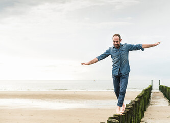 Carefree man with arms outstretched walking on wooden post at beach