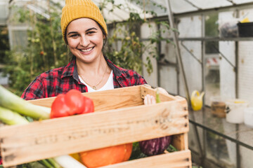 Smiling woman carrying crate with vegetables while standing against greenhouse