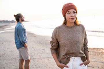 Beautiful young woman standing with hands in pockets against boyfriend at beach during sunset