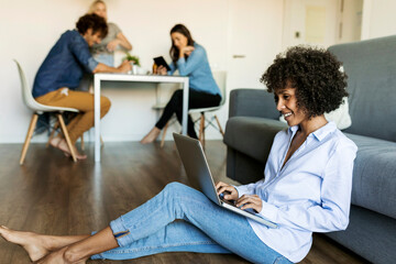 Smiling woman sitting on floor using laptop with friends in background