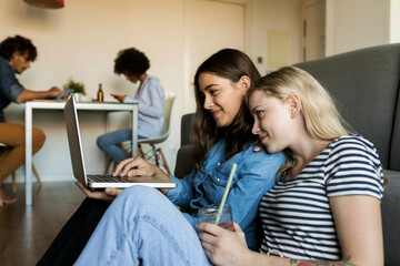 Two smiling young women sitting on floor sharing laptop with friends in background