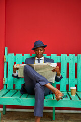 Young businessman wearing old-fashioned suit and hat sitting on a green bench reading the newspaper