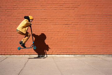 Boy performing stunt with push scooter on sidewalk against brick wall during sunny day - obrazy, fototapety, plakaty
