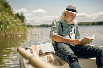 Portrait of content senior man sitting in rowing boat on a lake reading a book