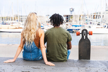 Spain, Barcelona, back view of multicultural young couple sitting side by side with longboard at marina