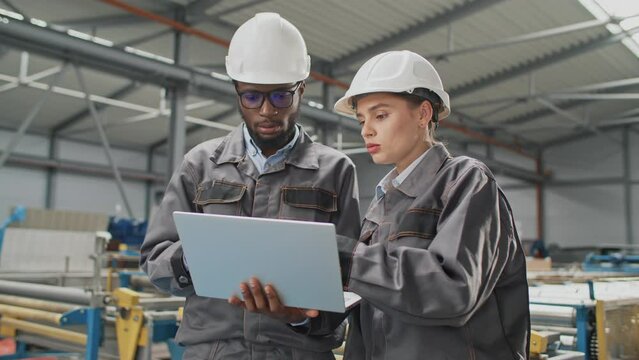 Two mixed-raced workers using laptop while standing in factory. Busy engineers looking for breakdown in production machine on technology device. Hardworking people speaking while working together.